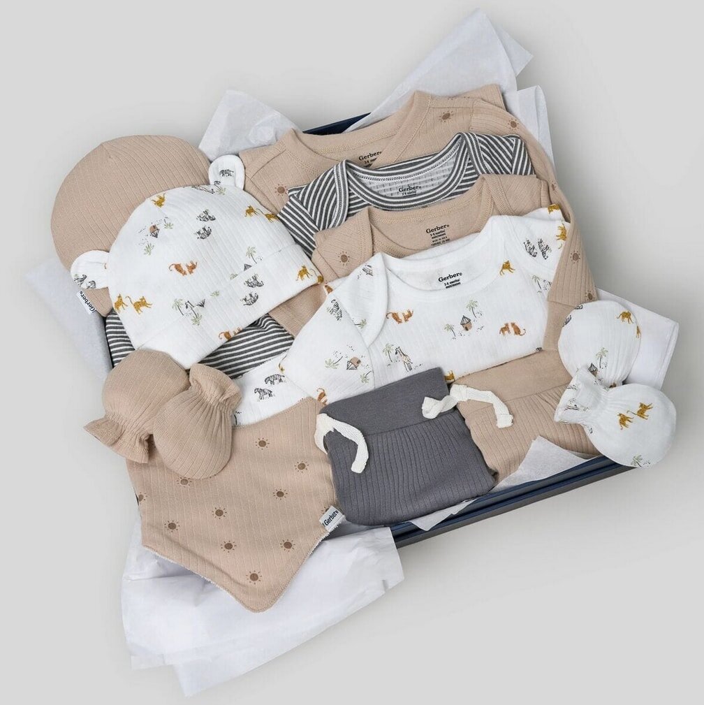 A neatly arranged baby gift set featuring soft beige, white, and gray clothing with animal prints, including onesies, a hat, mittens, and a bib.