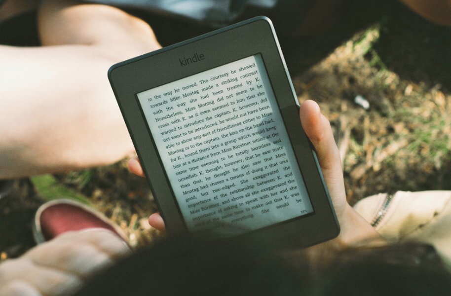 Person reading a Kindle outdoors. The e-reader displays text as they sit on grass with sunlight filtering through. A relaxed, peaceful scene.