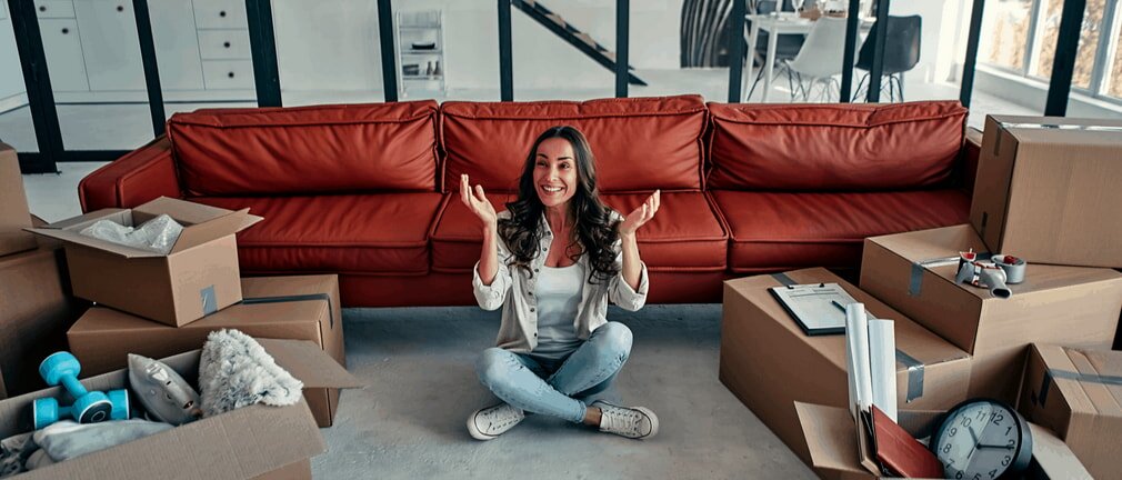 A cheerful woman sits cross-legged on the floor, surrounded by moving boxes. Behind her is a red sofa in a bright room, conveying excitement and a fresh start.