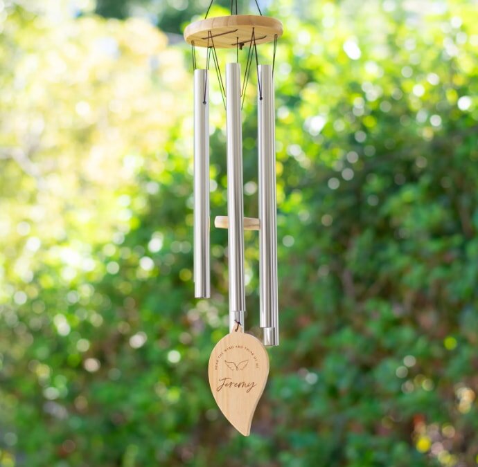 A wooden wind chime with silver tubes hangs outdoors against a blurred, leafy background. A heart-shaped pendant reads "Jeremy" with a butterfly design.