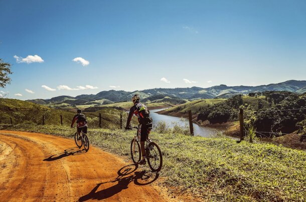 Two cyclists pedal along a rural dirt road, framed by green fields and a clear blue sky.