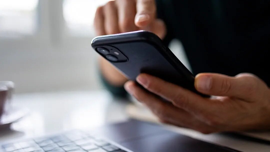 A person sitting at a desk, holding a cell phone and looking at the screen with a focused expression.
