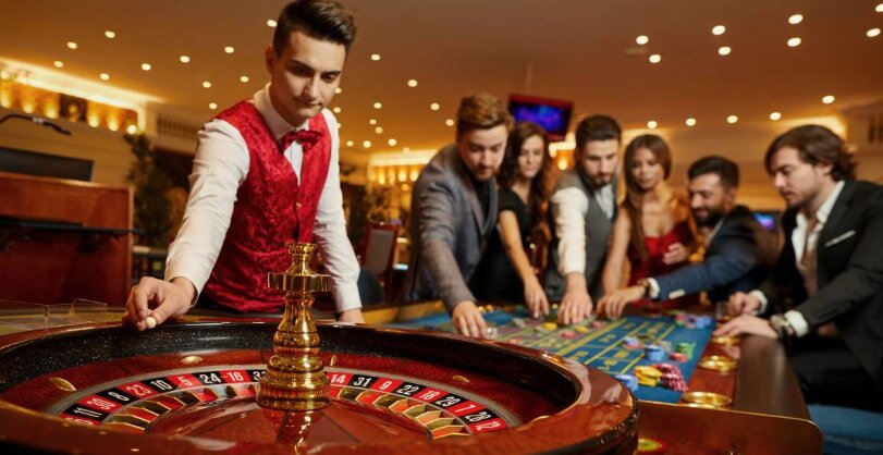 Casino scene with a roulette wheel in the foreground and a dealer in a red vest spinning. A group of people eagerly place bets at the table.