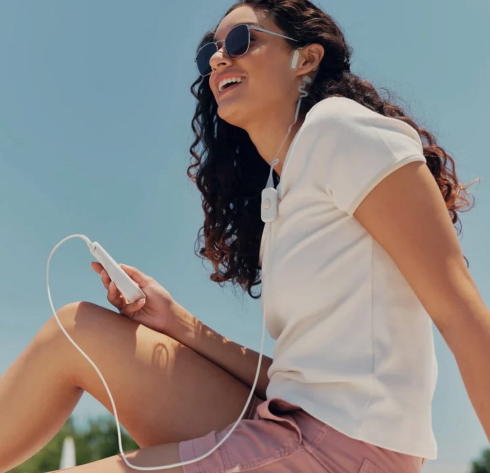 A woman sitting on a bench, wearing headphones, appears to be enjoying music or a podcast in a serene setting.