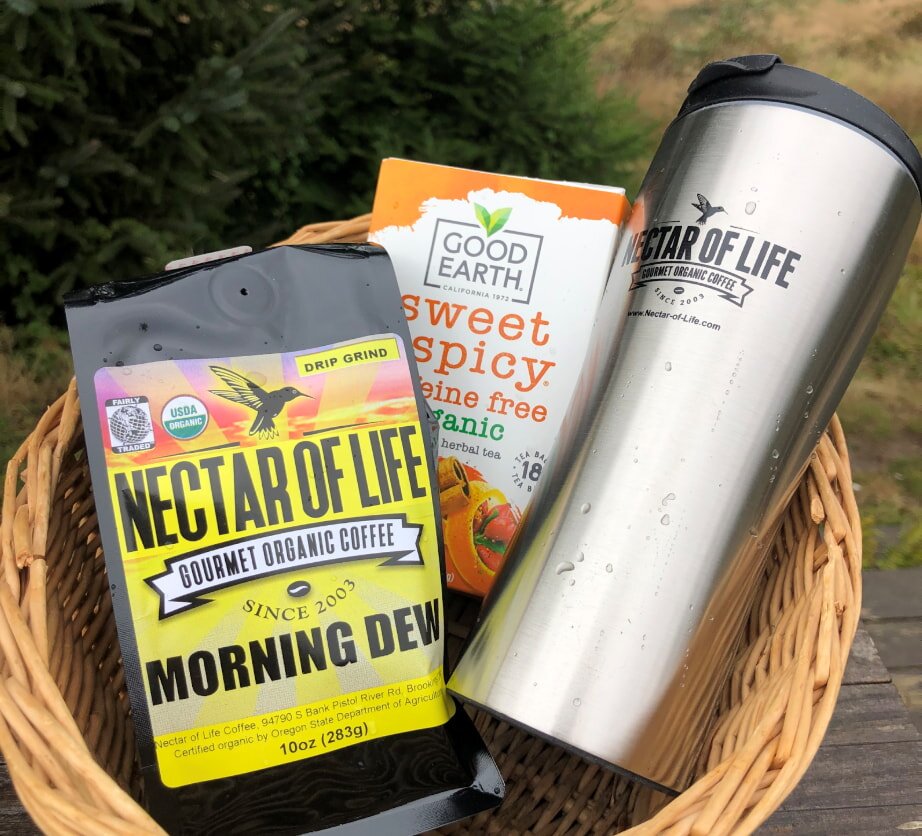 A wicker basket holds a bag of Nectar of Life organic coffee, a box of Good Earth sweet and spicy tea, and a stainless steel travel mug.