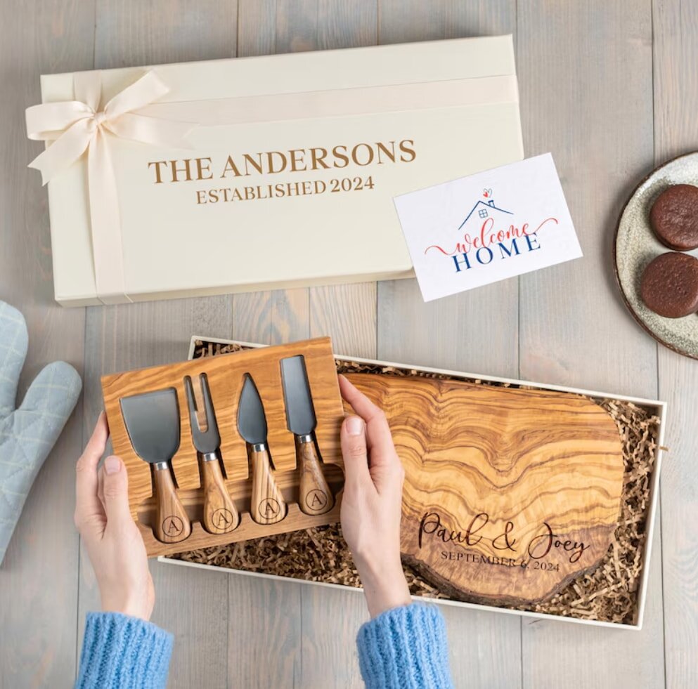 Hands hold a wooden cheese knife set inside an open gift box. Engraved cutting board reads "Paul & Joey, September 2024." Nearby, "Welcome Home" card and cookies. Elegant and inviting tone.