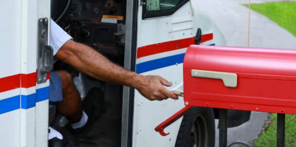 A postal worker in a white and blue mail truck leans out to deliver letters into a red mailbox. The scene conveys a routine, everyday task.