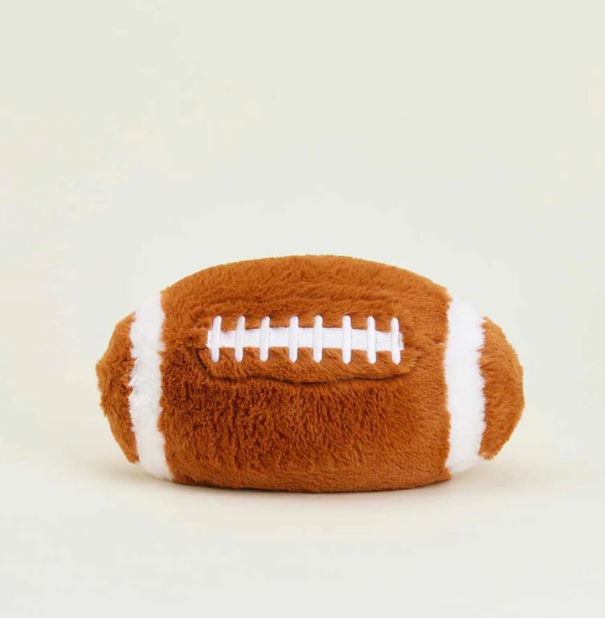 A plush football toy with brown fur and white laces, creating a soft, playful look. It sits on a light, neutral backdrop, evoking a cozy feel.