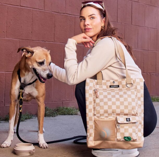 A woman kneels beside a dog and a bag, smiling as she interacts with the pet in a friendly outdoor setting.