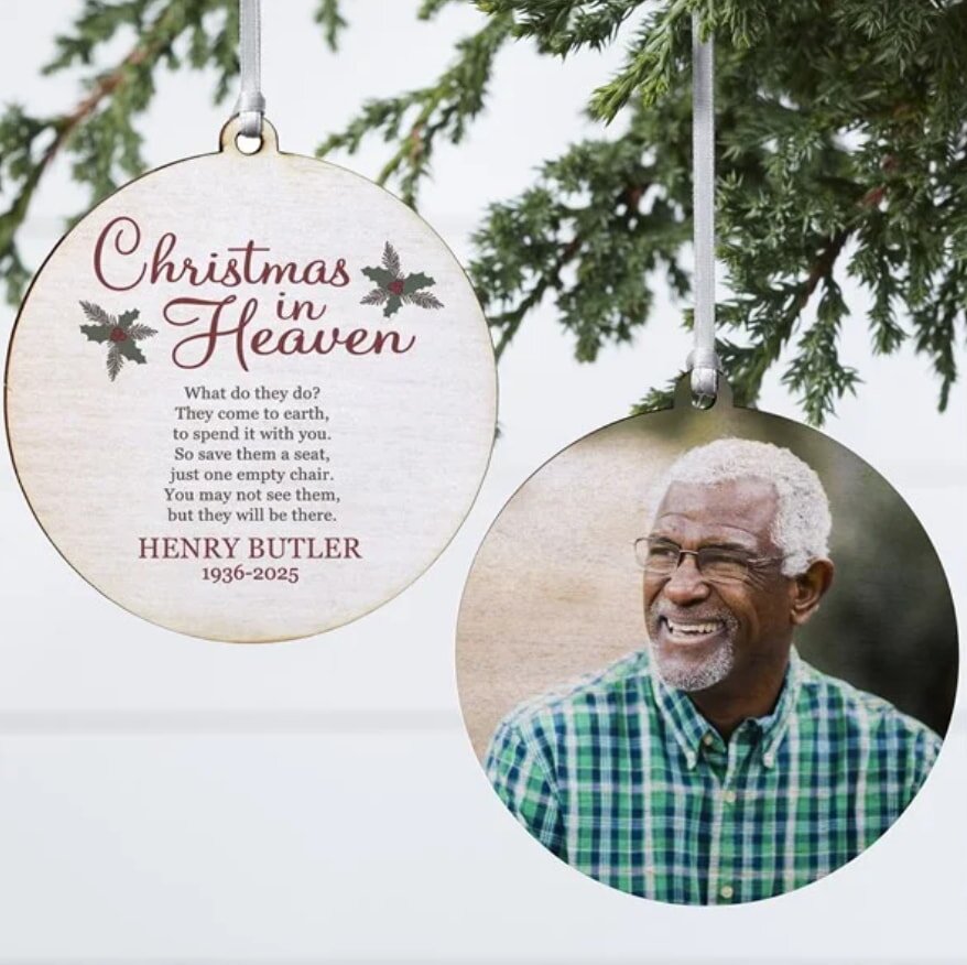 Two round ornaments hang from a pine branch. One features a smiling older man in a plaid shirt. The other has text about "Christmas in Heaven."