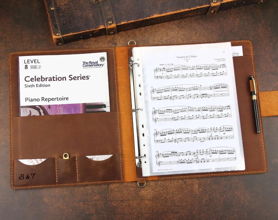 Leather music binder with piano sheet music on the right and a "Celebration Series" book in the left pocket. Pen placed in a side loop.