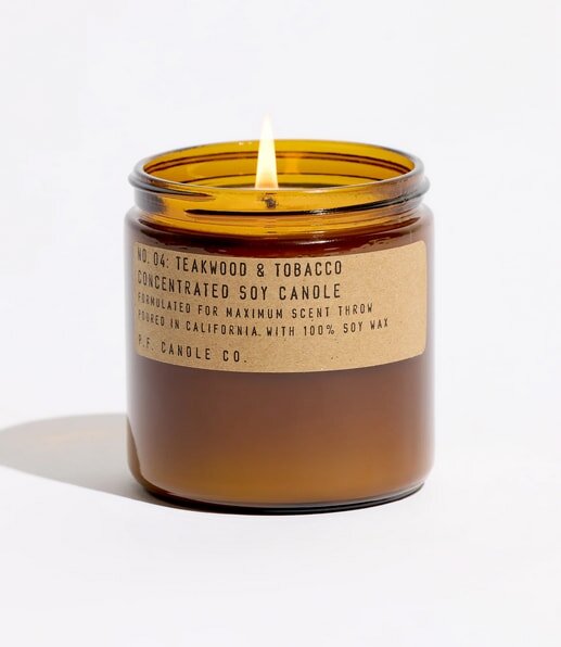 Brown jar candle labeled "Teakwood & Tobacco," lit with a bright flame, casting a soft shadow on a white background.