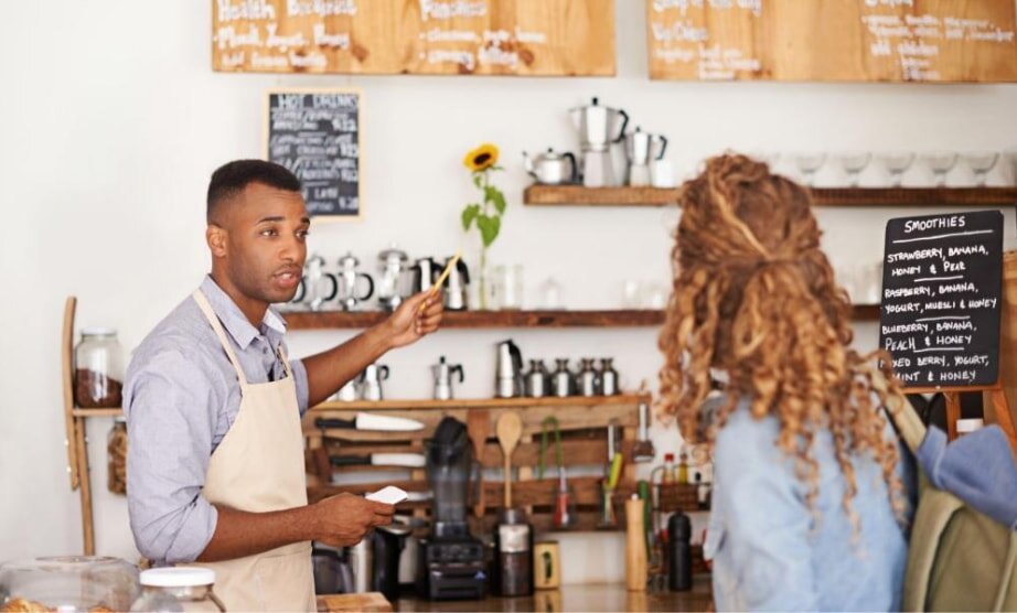 A barista in an apron gestures at a distant menu in a cozy café, assisting a customer. Shelves with mugs and a sunflower create a welcoming atmosphere.