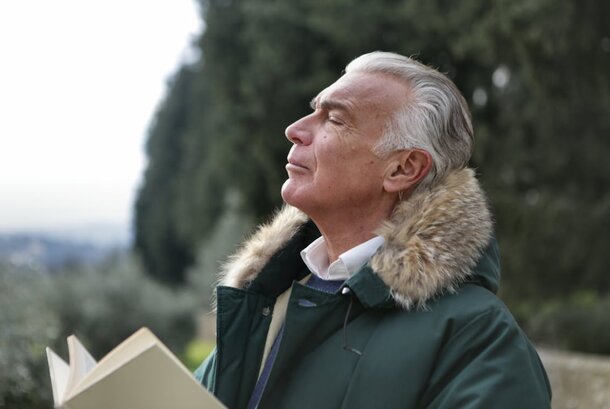 An older man in a green jacket holds a book, smiling as he stands in a cozy reading nook.