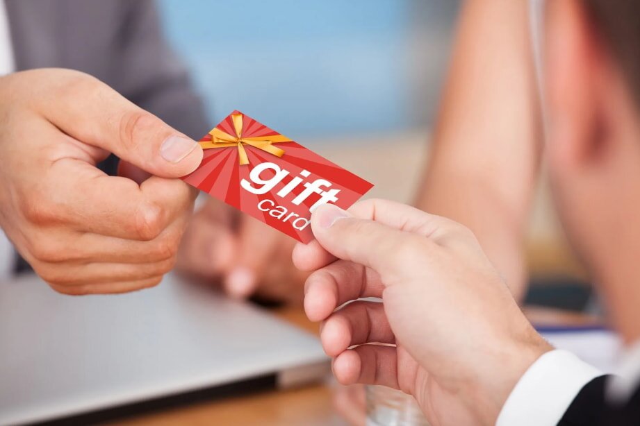 Two hands exchange a red gift card with a bow design in an office setting, creating a gesture of giving and warmth. A laptop is visible in the background.