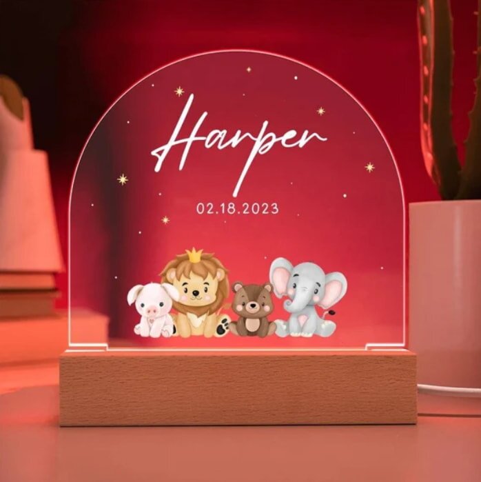 Clear acrylic night light display with a wooden base, featuring cute cartoon animals and the name "Harper" in white script above the date "02.18.2023" on a red background.