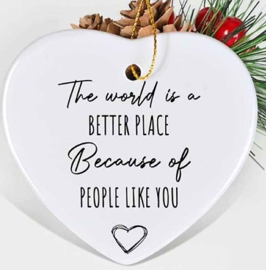 White heart-shaped ornament with black text: "The world is a better place because of people like you." Decorated with pine cones, red berries, and green sprigs.