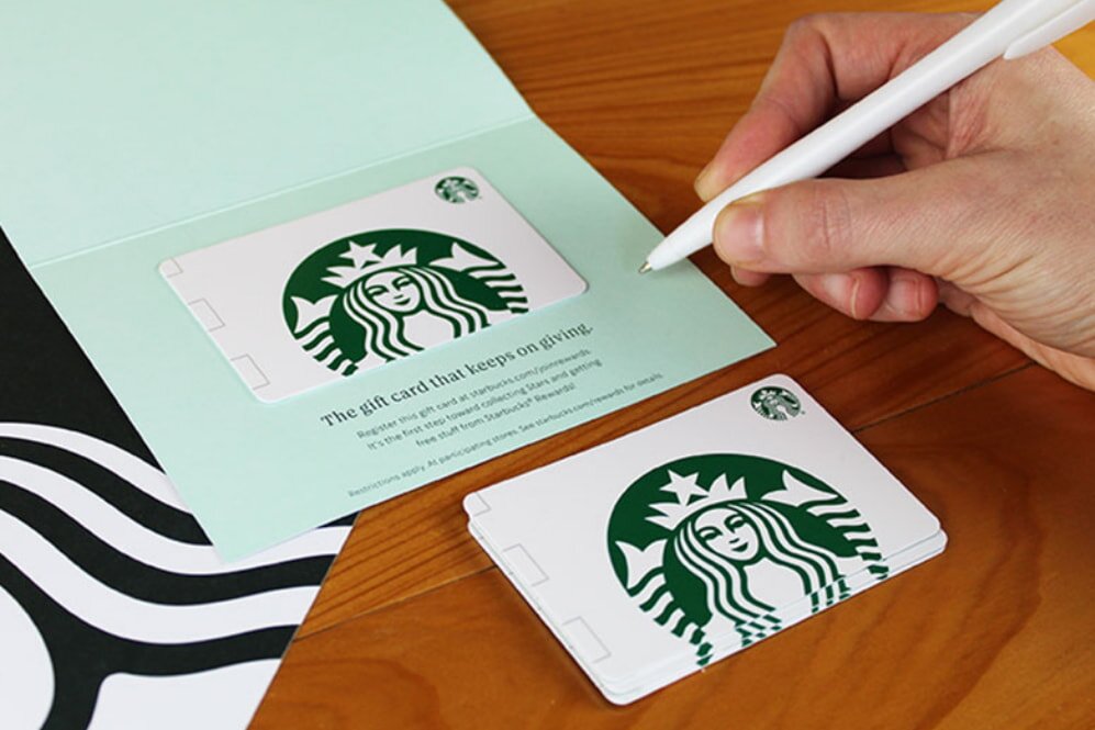 A hand holds a pen, writing on a green card displaying a Starbucks gift card with the logo. Another gift card lies beside it on a wooden table.