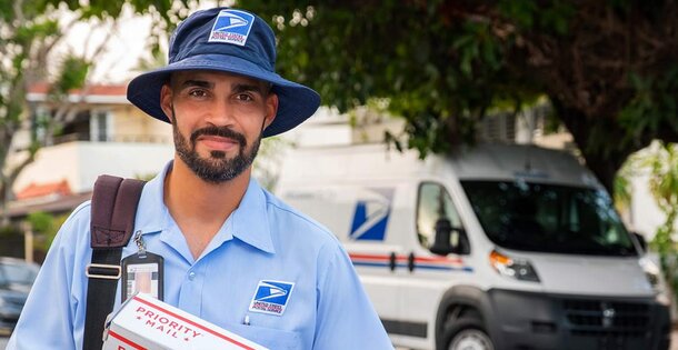 Postal worker in a blue uniform and bucket hat holds a priority mail package, smiling warmly. A postal van is visible in the background, under trees.