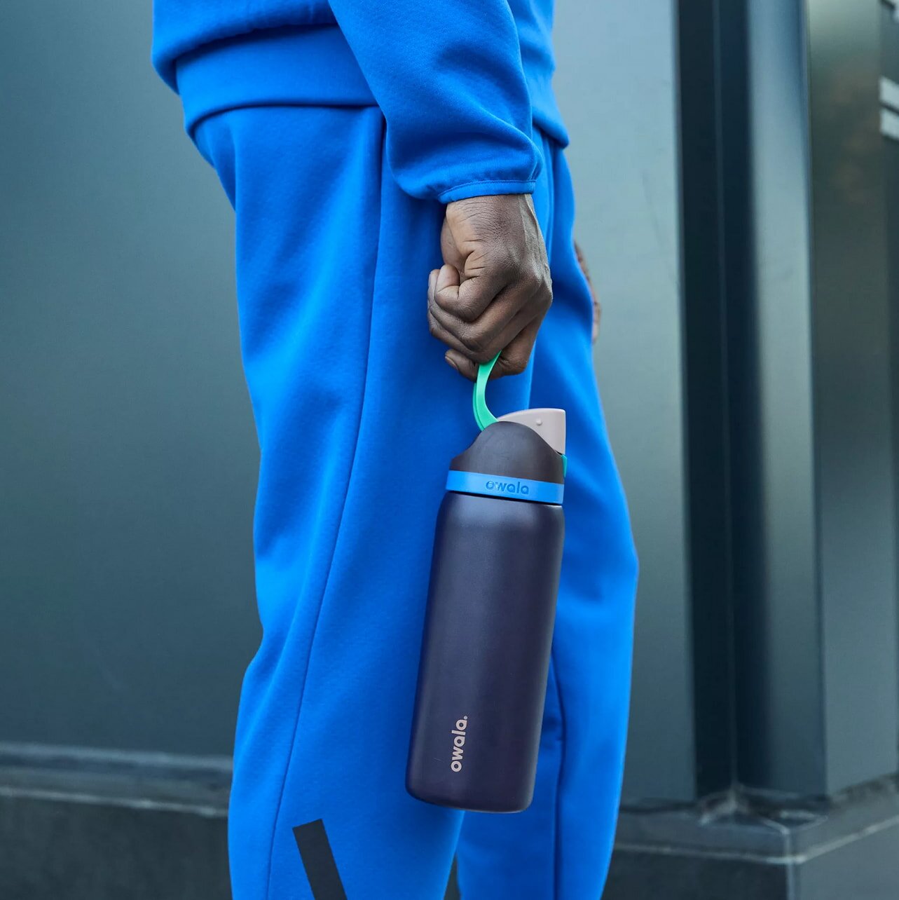 A man wearing blue sweatpants and a blue jacket holds a water bottle, standing in a casual pose.