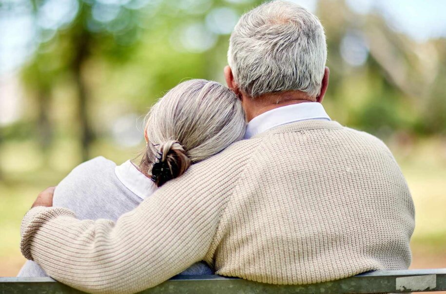 Elderly couple sitting on a bench, woman resting her head on man's shoulder. They're dressed in beige sweaters, enjoying a peaceful day in a park.