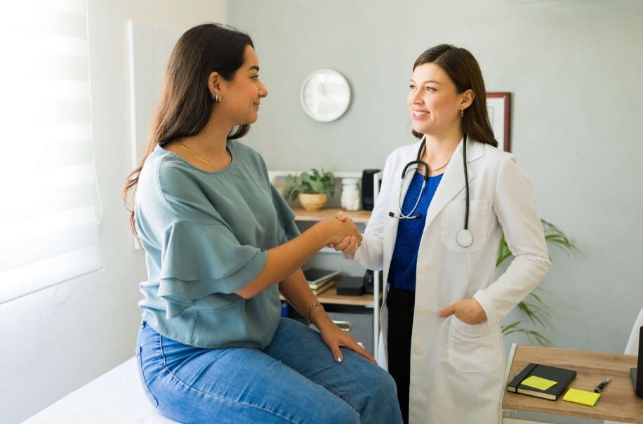 A doctor and patient warmly shake hands in a medical office. The patient sits on an exam table, while the doctor in a white coat smiles reassuringly.