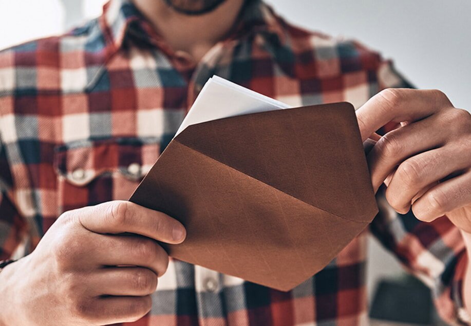 A person in a plaid shirt opens a brown envelope, revealing a white paper inside. The scene conveys anticipation or curiosity.