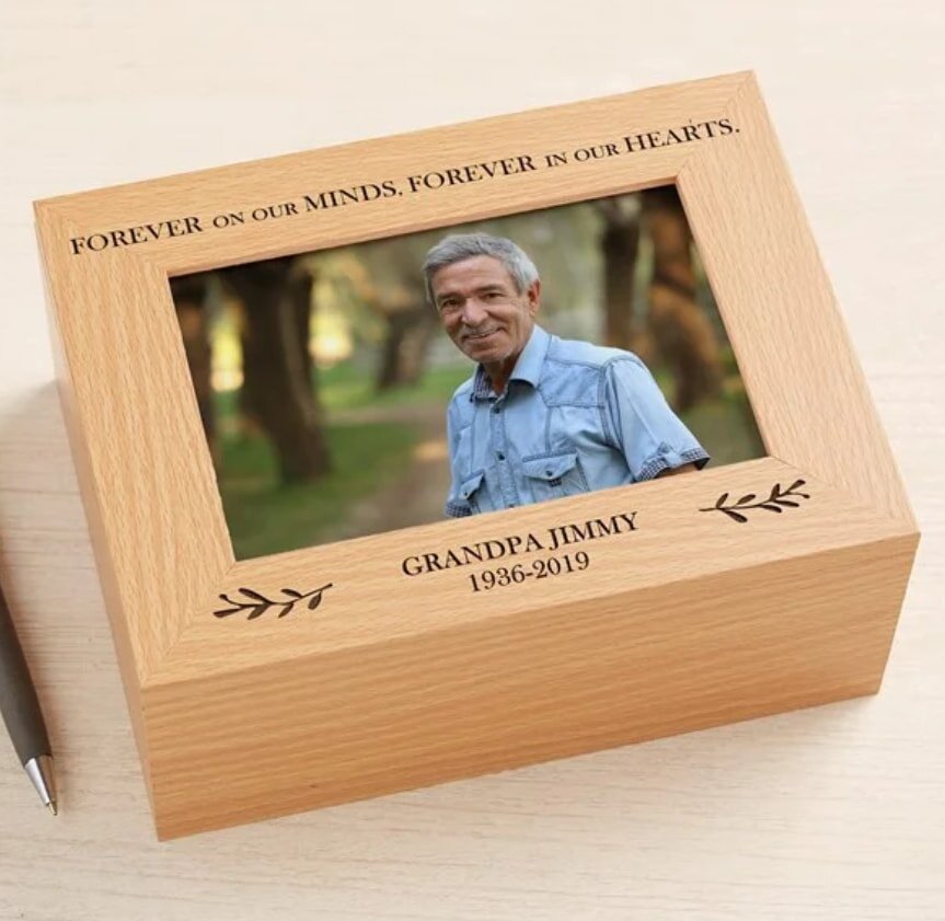Wooden memory box with a photo of an older man smiling in a park. Text: "Forever on our minds. Forever in our hearts. Grandpa Jimmy 1936–2019". Serene atmosphere.