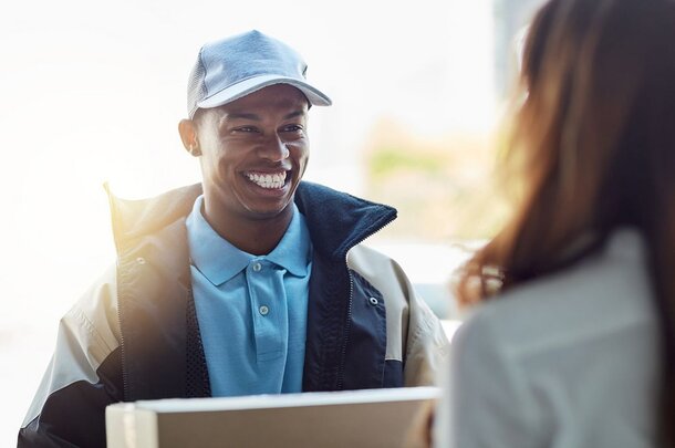 A smiling delivery person in a cap and uniform jacket hands a package to a woman. The scene is bright and friendly, conveying a positive tone.