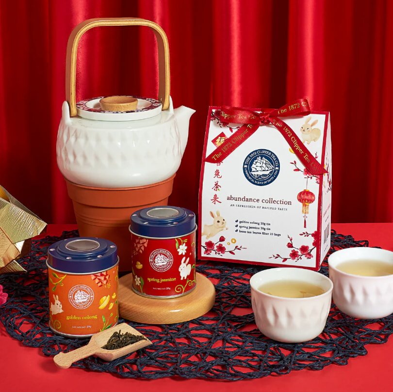 Tea gift set with a white teapot on a terracotta warmer, two tea tins, and two tea cups on a red table. A festive bag and vibrant red curtain backdrop.