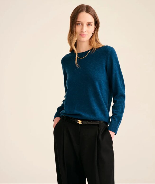 A person wearing a blue sweater and black pants stands against a neutral background with a slight smile. They exude a relaxed, confident tone.