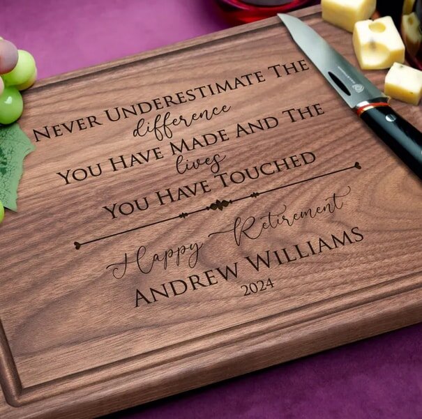 Wooden board engraved with retirement message for Andrew Williams 2024, surrounded by grapes, cheese, and a knife.