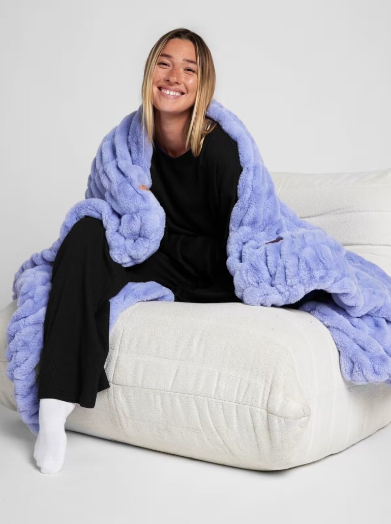 Smiling woman wrapped in a plush, blue blanket sits on a white sofa. She wears black clothes and white socks, exuding comfort and warmth.
