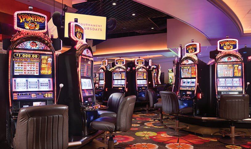 A casino interior featuring rows of slot machines and chairs arranged in the center for players.