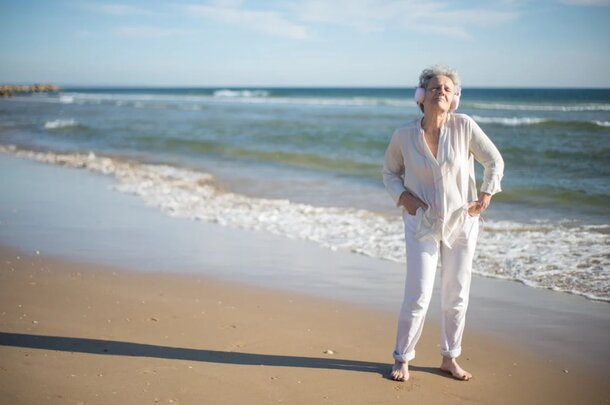 An older woman stands on the beach, gazing at the ocean waves under a clear blue sky.