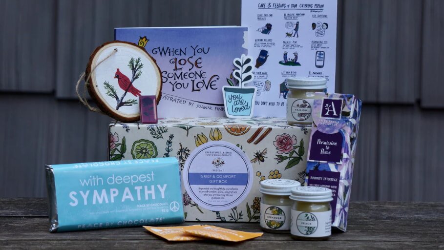 Gift box with floral design labeled "Grief & Comfort" contains a sympathy card, chocolate bar, candles, tea, a book, and a plaque. It conveys warmth and solace.