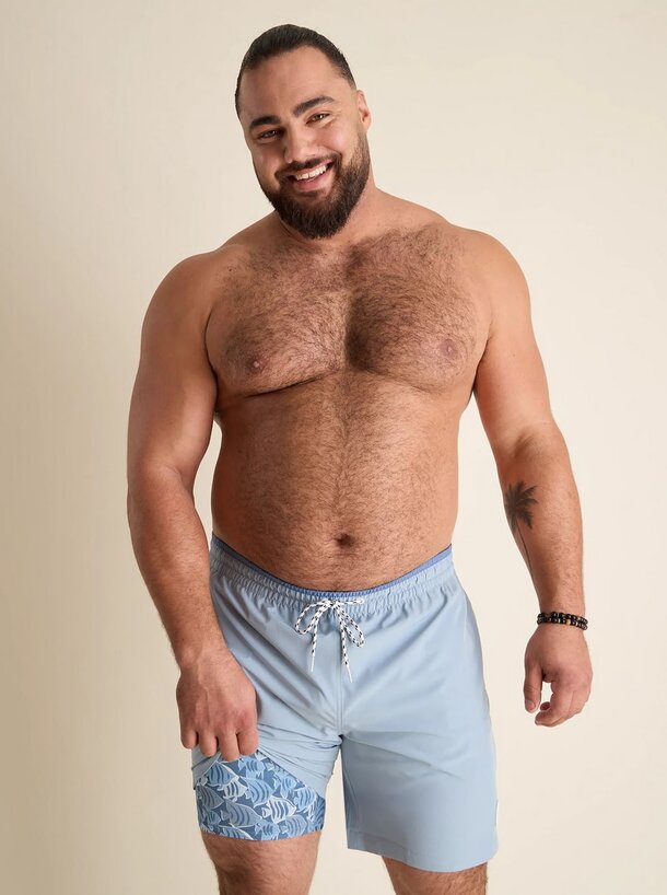 A shirtless man with a beard wearing blue swim shorts stands confidently, showcasing a relaxed beach vibe.
