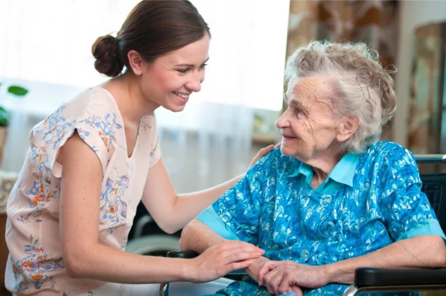 A young woman smiles warmly at an elderly woman in a wheelchair. They hold hands in a comforting, joyful interaction. The room is softly lit and homey.