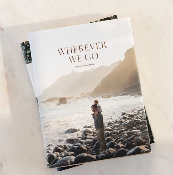 A book cover titled "Wherever We Go" shows a person carrying a child on a rocky beach. Ocean waves and misty mountains create a serene, warm backdrop.