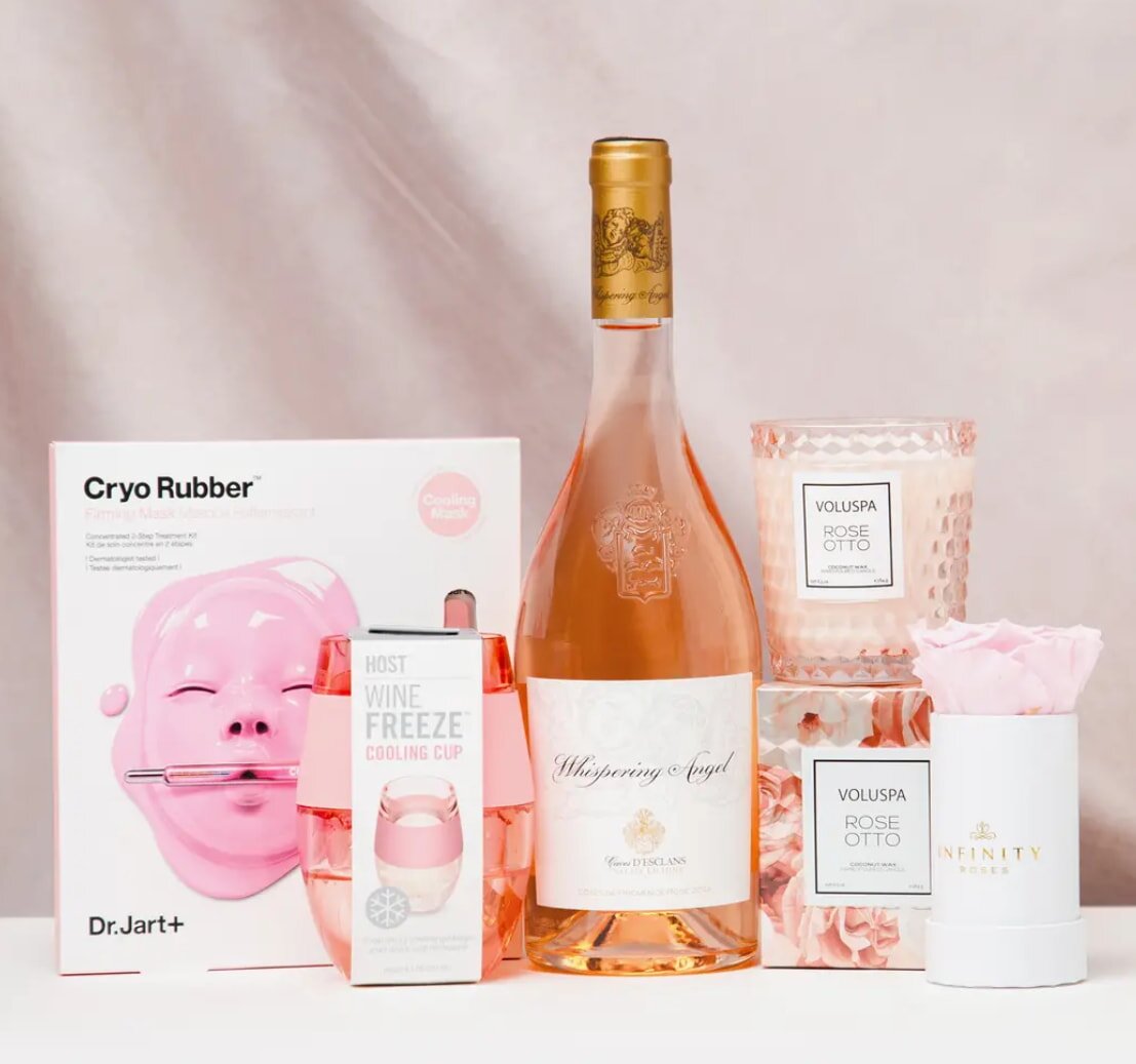A pink wine bottle, candle, face mask, and gift box arranged together on a neutral background.