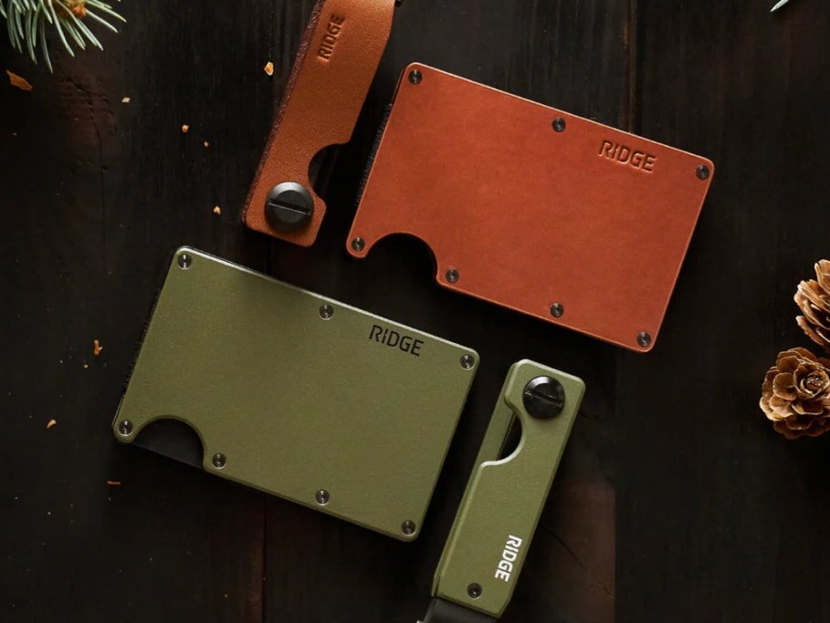 Stylish wallets and knives on dark wood, featuring olive green and brown leather. Pinecones and greenery add a festive, rustic feel.