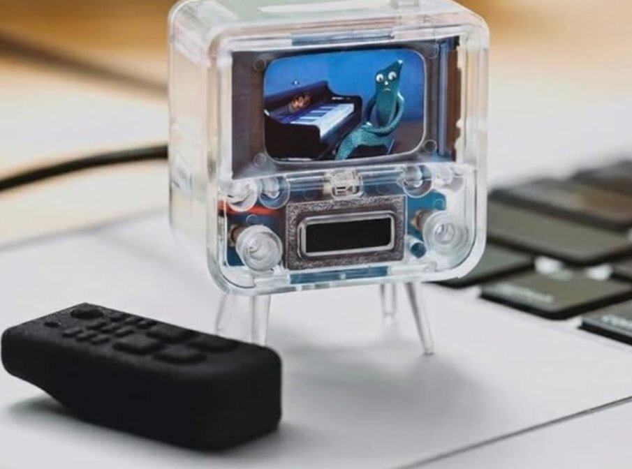 A small retro-style TV with a clear case displays a cartoon character playing a piano. A black remote is nearby on a laptop keyboard, evoking nostalgia.