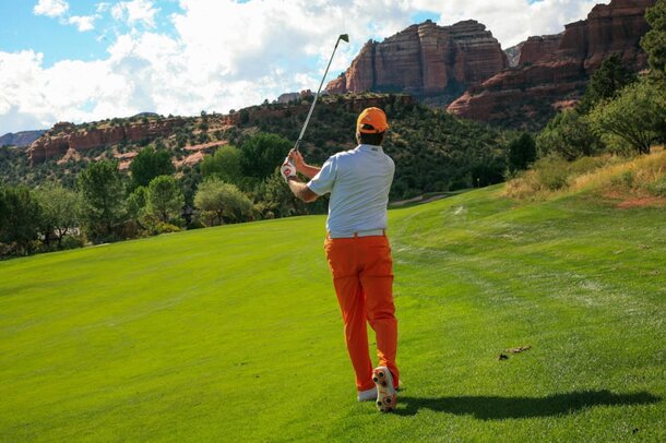 A man in an orange shirt and pants swings a golf club on a sunny course.