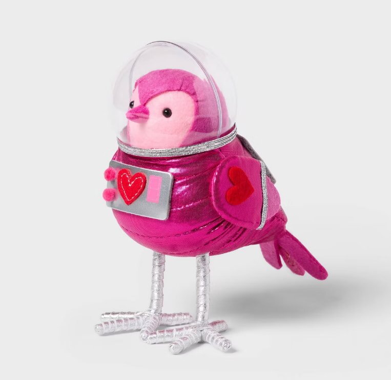 Plush pink bird toy in a metallic purple spacesuit with heart designs, wearing a clear helmet. It stands on curly silver legs, conveying a playful tone.