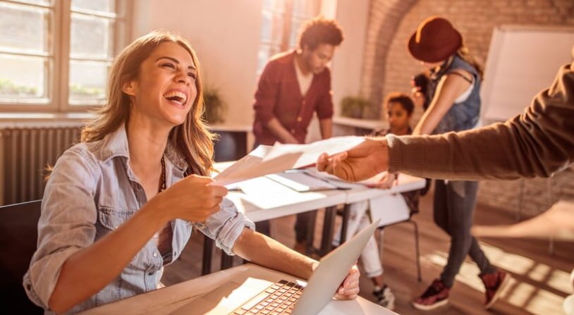 A smiling woman holds a piece of paper, conveying a sense of joy and accomplishment.