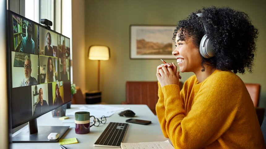 A woman with headphones sits at a desk, engaged in a video call on her computer screen.