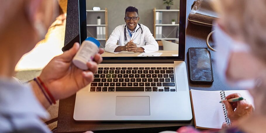 An elderly couple engages in a video call with a smiling doctor on a laptop. The woman takes notes while the man holds a medication bottle, suggesting a telehealth consultation.