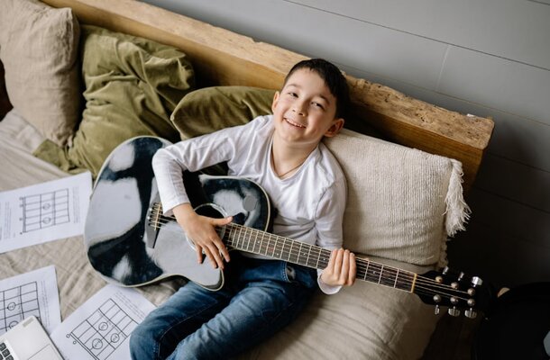 A boy sits on a bed, holding a guitar, looking focused as he prepares to play music.