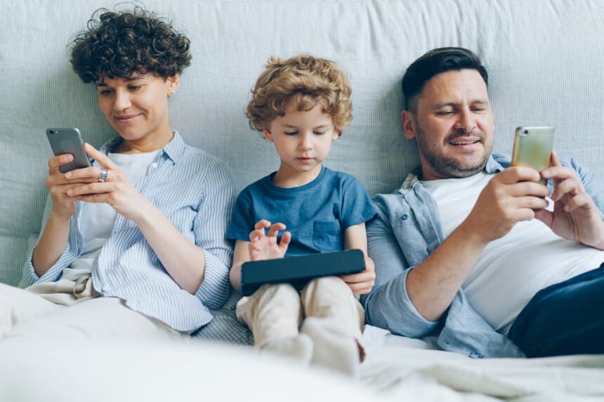 A family of four sitting on a bed, each using their smartphones, engaged in their own activities.