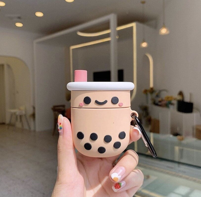 A hand with colorful nail art holds a cute keychain shaped like a smiling boba tea cup in a modern, softly lit cafe with minimalistic decor.